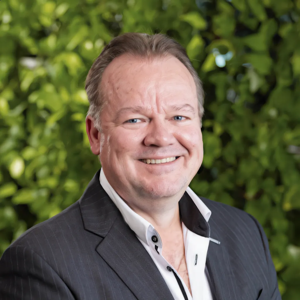 Professional headshot of Derrick Vickers with a blurred background of green foliage.